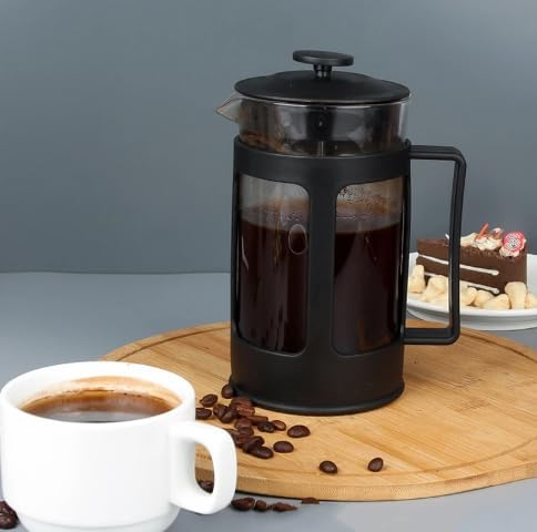 Black French press filled with coffee on a wooden board with a cup of coffee and scattered coffee beans.