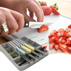 Person cutting strawberries with a knife on a cutting board, surrounded by a set of colorful kitchen utensils.