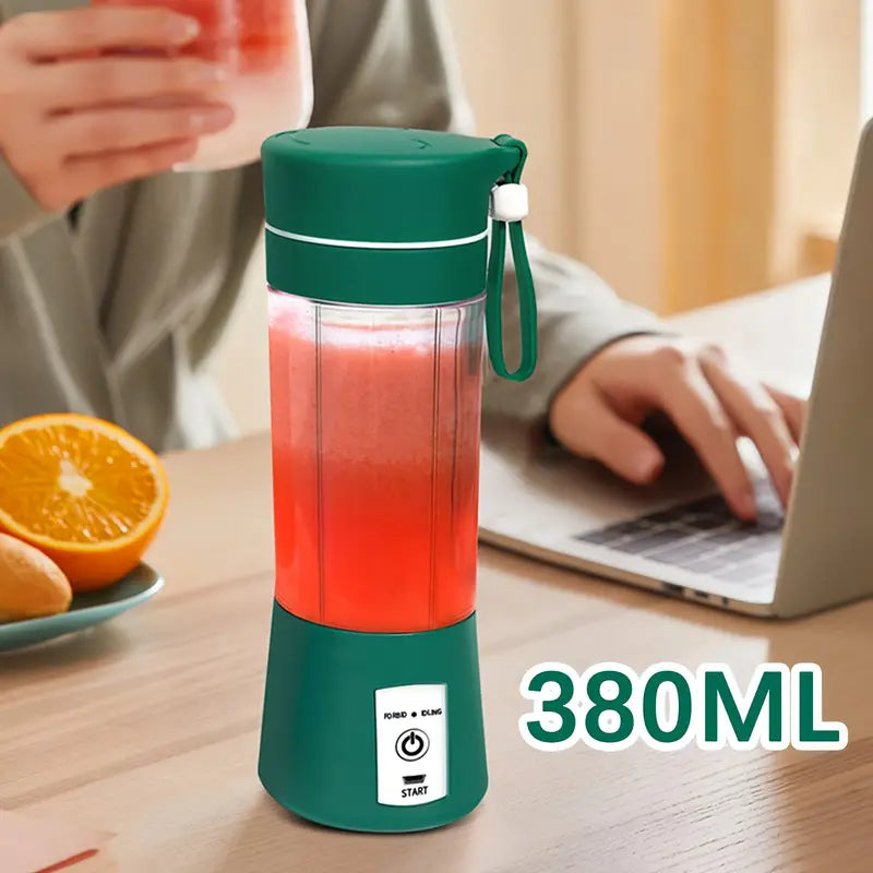 Green portable blender with red juice on a table next to a person using a laptop and an orange.
